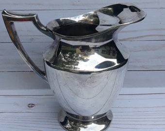 May include: A silver pitcher with a wide, rounded spout and a handle. The pitcher has a polished, reflective surface.