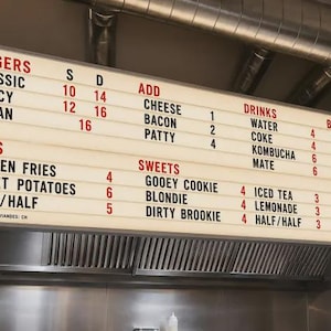 May include: A rectangular, illuminated menu board with a white background and black text. It lists food and drink categories including Burgers, Sides, Add, Sweets, Drinks, and Beers, with corresponding prices. The text 'MADE WITH LOVE' is in the bottom right corner.