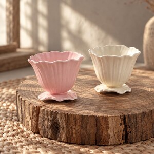 May include: Two small ceramic cups with a fluted, petal-like design. One cup is light pink and the other is off-white. Each cup features a scalloped rim and a base shaped like a small flower, providing a stable foundation for the bowl-shaped vessel.