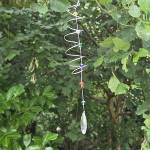 May include: A silver-colored metal spiral wind chime with colorful beads and a clear crystal pendant. The beads are yellow, clear, green, blue, and red. The wind chime hangs from a tree branch against a backdrop of green foliage.