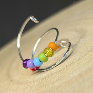 Könnte beinhalten: Ein silberfarbener, spiralförmiger Ring mit einer Reihe farbiger Perlen. Die Perlen sind in einem Regenbogenmuster angeordnet, mit roten, violetten, blauen, grünen, gelben und orangenen Perlen. Der Ring ist für das Ohr gedacht.