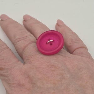May include: A bright pink button ring with a silver metal ring. The ring is worn on a person's finger.