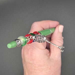 Könnte beinhalten: Ein grüner Stift mit dekorativen Perlen und einem silbernen Anhänger. Der Stift hat eine grüne Spitze und ist mit roten, grünen und silbernen Perlen verziert. Ein silberner Anhänger hängt am Stift und verleiht ihm einen Hauch von Eleganz. Der Stift wird in einer Hand gehalten.