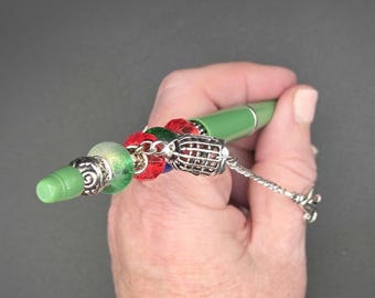 May include: A green pen with decorative beads and a silver charm. The pen has a green tip and is adorned with red, green, and silver beads. A silver charm hangs from the pen, adding a touch of elegance. The pen is held in a hand.