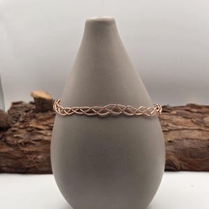 May include: A rose gold bracelet with a woven design is displayed on a gray, teardrop-shaped vase. The bracelet's intricate pattern is highlighted against the vase's smooth surface. The background includes a piece of wood, adding a natural element to the composition.