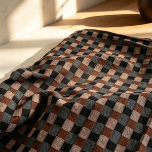 May include: A textile fabric featuring a repeating checkered pattern in shades of deep brown, black, and muted blue. The material displays a visible woven texture with a grid-like design, characterized by soft folds and creases across the surface.