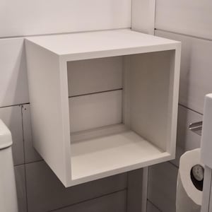 May include: A white, square, open-front wall-mounted storage cube with a smooth finish. The unit features a hollow interior space and is constructed with clean, straight edges, providing a simple, box-like structure for organizing items.