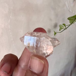 Könnte beinhalten: Eine klare, facettierte Quarzkristallgruppe mit sichtbaren natürlichen Streifen und inneren Rissen. Das Mineralexemplar weist eine spitze Endung und ein transparentes, glasartiges Aussehen auf, das Licht auf seinen geometrischen Oberflächen reflektiert.