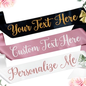May include: Three satin sashes with different colors and text. The first sash is black with gold glitter text that says "Your Text Here". The second sash is pink with white text that says "Custom Text Here". The third sash is white with rose gold glitter text that says "Personalize Me".