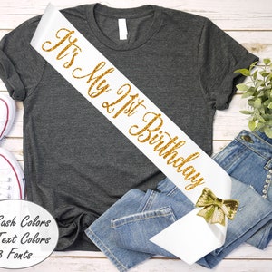 May include: White satin sash with gold glitter text that reads "It's My 21st Birthday". A gold glitter bow is attached to the sash.