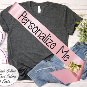 May include: Pink satin sash with a gold bow and the text "Personalize Me" in black. The sash is displayed on a gray t-shirt, blue jeans, and a white wooden surface.