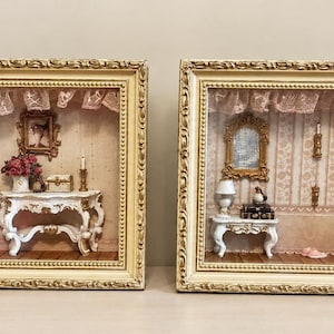 May include: Two rectangular shadow boxes with ornate gold frames. The left box displays a miniature room with a white tufted armchair and a console table. The right box features a miniature bedroom with a white bed, a side table, and a mirror.