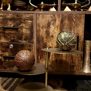 May include: A tiered metal display stand with three circular shelves in a matte finish. A brown woven sphere sits on the lower shelf, while a green and gold textured sphere with a segmented pattern rests on the upper shelf.