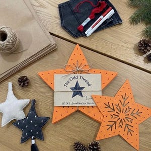May include: A set of three orange star-shaped stencils with snowflake cutouts, labelled The Odd Thread, accompanied by a white fabric star, a dark grey star ornament with white stars, a ball of twine, red embroidery thread, and two pens.