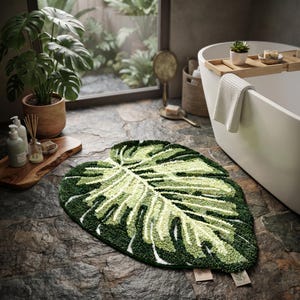 May include: A tufted bath mat shaped like a large monstera leaf, featuring a textured pile in various shades of dark green, light green, and cream, with detailed vein patterns mimicking the natural structure of a tropical plant leaf.