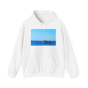 Puede incluir: Una sudadera blanca de manga larga con capucha y un estampado gráfico rectangular en el pecho. El estampado muestra un barco oscuro flotando en un mar azul profundo bajo un cielo azul claro, con una costa visible en el horizonte.