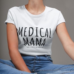 May include: A white short-sleeved t-shirt featuring the black text MEDICAL MAMA and the smaller text YOU CAN'T SCARE US printed on the front. The shirt has a crew neckline and is paired with blue denim jeans that have frayed hem details.