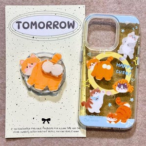 Könnte beinhalten: Eine transparente Handyhülle und ein runder Handyhalter mit orange-weißen Katzenillustrationen. Die Hülle zeigt mehrere Katzen, Sterne, Herzen und den Text "HAPPY CUTE". Die Verpackungskarte enthält das Wort "TOMORROW" am oberen Rand.