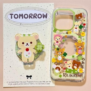 Könnte beinhalten: Eine transparente Handyhülle und ein runder Handyhalter mit Rilakkuma-Figuren. Die Hülle hat einen grünen Rand und verschiedene Bärenillustrationen mit Kleeblattmotiven. Der Handyhalter zeigt einen hellgelben Bären mit rotem Punkt.
