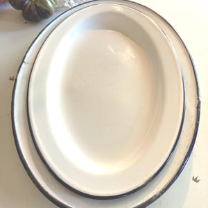 May include: Two stacked oval enamel serving platters with a glossy off-white finish and a dark blue rim. The metal surfaces show visible signs of wear, including light surface scratches and minor chips along the edges of the dark blue trim.