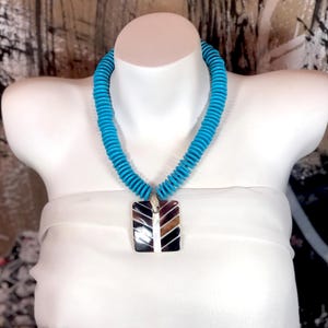 Puede incluir: Un collar de cuentas turquesa con un colgante cuadrado que presenta un diseño geométrico en marrón, negro y blanco.