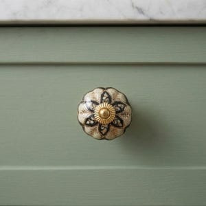 May include: A round ceramic cabinet knob featuring a scalloped edge and a floral pattern in shades of cream, black, and gold. The center of the knob is accented with a small, raised, circular gold-toned metal detail.