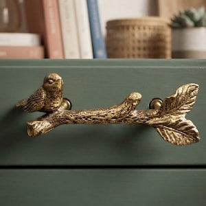 May include: A gold-toned metal drawer pull shaped like a textured tree branch with a small bird perched on the left side and a leaf at the right end. The hardware is mounted horizontally onto a flat, matte sage green surface with two circular attachment points.