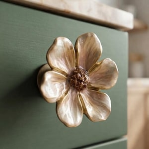 May include: A flower-shaped cabinet knob with a metallic gold finish and soft pinkish-white petals. The center of the flower features a textured, dark brown stamen detail. The knob is mounted onto a flat, matte olive green drawer front surface.