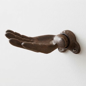 May include: A wall-mounted hook shaped like an open human hand, crafted from dark brown, textured cast metal. The hand is positioned with the palm facing upward, extending outward from a circular base plate secured by two visible screws.