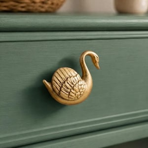 May include: A gold-toned metal drawer knob shaped like a swan with detailed wing feathers. The swan is mounted on a flat, matte sage green drawer front. The knob features a curved neck and a textured body, providing a functional handle for furniture.