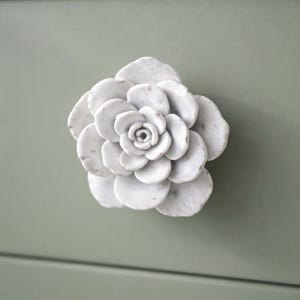 May include: A white, multi-layered flower-shaped cabinet knob with a smooth, stone-like texture and subtle grey veining. The knob features overlapping, rounded petals arranged in a circular pattern, creating a dimensional, sculptural appearance.