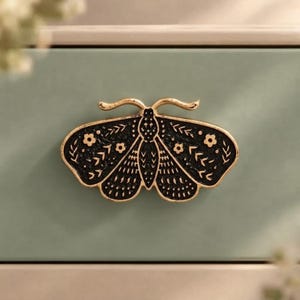 May include: A black and gold metal drawer knob shaped like a moth. The moth features intricate gold-colored etched patterns including small flowers, leaves, and geometric lines against a textured black background with gold edges.