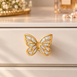 May include: A gold-toned butterfly-shaped drawer knob featuring inlaid iridescent white stone segments within its wings. The hardware is mounted onto a smooth, light-colored wooden drawer front, showcasing a symmetrical design with a metallic finish.