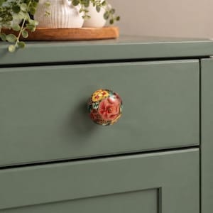 May include: A round ceramic drawer knob featuring a detailed floral pattern with vibrant red, yellow, and green colors. The knob is mounted on a solid sage green drawer front, providing a clear view of the intricate painted botanical design.