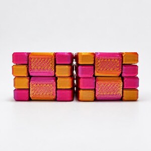 K&ouml;nnte beinhalten: Zwei rechteckige Bl&ouml;cke mit einem Gitter aus kleineren, erhabenen Quadraten. Die Oberfl&auml;che zeigt einen lebendigen Farbverlauf aus metallischem Pink und Orange, wobei auf jedem einzelnen quadratischen Teil eine 3D-Druckstruktur erkennbar ist.