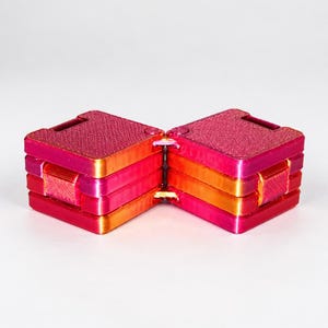 K&ouml;nnte beinhalten: Ein quadratisches Fidget-Spielzeug mit zwei Abschnitten in einem lebendigen Farbverlauf aus Magenta, Pink und Orange. Das Objekt besteht aus zwei miteinander verbundenen, geschichteten Bl&ouml;cken mit einer texturierten 3D-Druck-Oberfl&auml;che, die durch ein zentrales Scharnier verbunden sind.