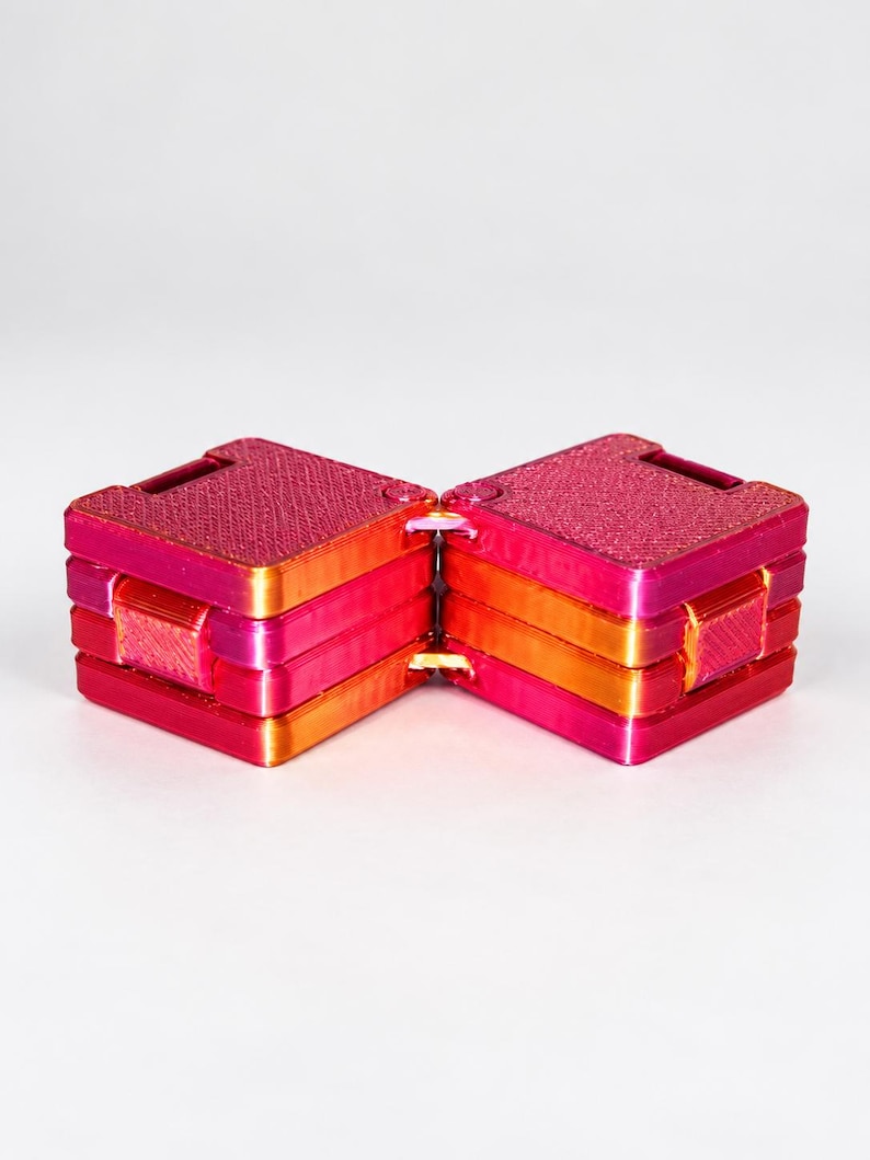 K&ouml;nnte beinhalten: Ein quadratisches Fidget-Spielzeug mit zwei Abschnitten in einem lebendigen Farbverlauf aus Magenta, Pink und Orange. Das Objekt besteht aus zwei miteinander verbundenen, geschichteten Bl&ouml;cken mit einer texturierten 3D-Druck-Oberfl&auml;che, die durch ein zentrales Scharnier verbunden sind.