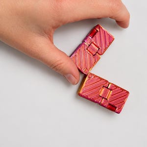 K&ouml;nnte beinhalten: Ein Fidget-Spielzeug bestehend aus zwei rechteckigen, klappbaren Segmenten mit einer metallisch schimmernden Oberfl&auml;che, die zwischen Pink, Magenta und Orange wechselt. Die Oberfl&auml;che weist ein diagonales Rillenmuster auf.