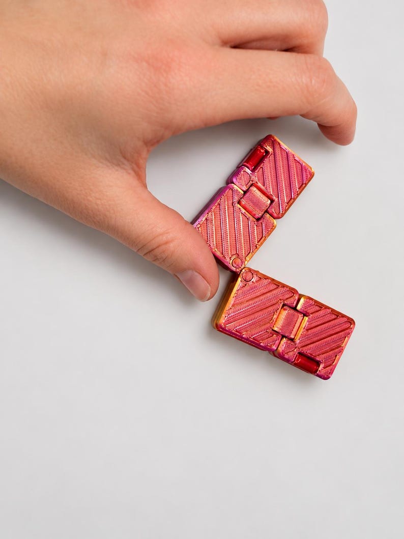 K&ouml;nnte beinhalten: Ein Fidget-Spielzeug bestehend aus zwei rechteckigen, klappbaren Segmenten mit einer metallisch schimmernden Oberfl&auml;che, die zwischen Pink, Magenta und Orange wechselt. Die Oberfl&auml;che weist ein diagonales Rillenmuster auf.