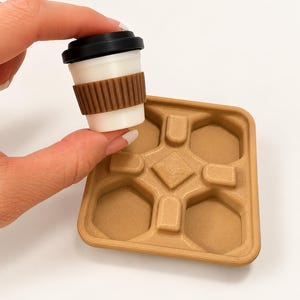 Könnte beinhalten: Ein weißer Miniatur-Kaffeebecher mit schwarzem Deckel und brauner gerippter Manschette, gehalten über einem quadratischen beigen Tablett mit vier sechseckigen Vertiefungen und einem strukturierten rautenförmigen Detail.