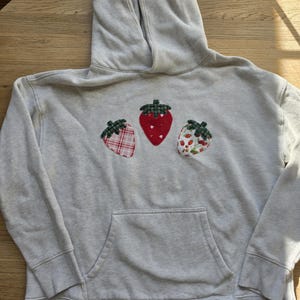 May include: A light gray hooded sweatshirt featuring three fabric strawberry appliqués on the chest. The center strawberry is solid red, while the left one has a red plaid pattern and the right one features a small red strawberry print.