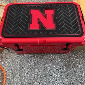 May include: A bright red rectangular cooler featuring a black textured top panel with a herringbone pattern. A large, bold red letter N is centred on the black panel. The cooler has black rubber latches on the front and a smooth, glossy finish.