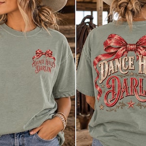 May include: A sage green short-sleeve t-shirt featuring a red plaid bow graphic and the text "DANCE HALL DARLIN'" on the front left chest and a larger version of the same graphic and text printed across the back of the garment.