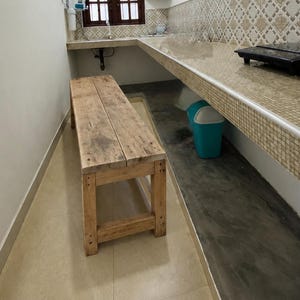 Könnte beinhalten: Eine rechteckige Holzbank mit natürlicher hellbrauner Oberfläche und sichtbarer Holzmaserung. Die Bank verfügt über eine flache Sitzfläche, die von vier stabilen Beinen getragen wird, welche durch horizontale Querstreben verbunden sind.