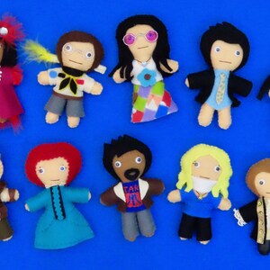 May include: A collection of handmade felt dolls, each depicting a different character. The dolls vary in color and design, with some wearing outfits and accessories. The dolls are arranged on a blue surface.