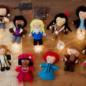 May include: A collection of handmade felt dolls, each depicting a different character. The dolls feature various outfits and hairstyles, with details like glasses, beards, and accessories. The dolls are arranged on a wooden surface with string lights.