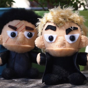 May include: Two plush toys, one with black hair and one with blonde hair, both wearing black jackets. The toys have painted-on facial features and are sitting on a wooden surface.