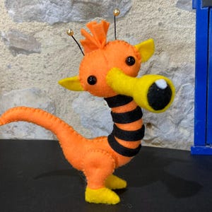 May include: A handmade orange felt creature with yellow accents, black stripes, and a large yellow eye-like feature. The whimsical design includes small antennae with gold beads and black button eyes. The creature stands on yellow feet.