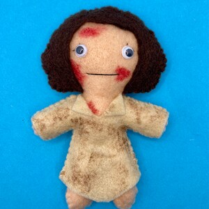 May include: A handmade felt doll with a light tan face, brown hair, and a light tan dress. The doll has red markings on its face and chest, and googly eyes. The doll is set against a blue background.