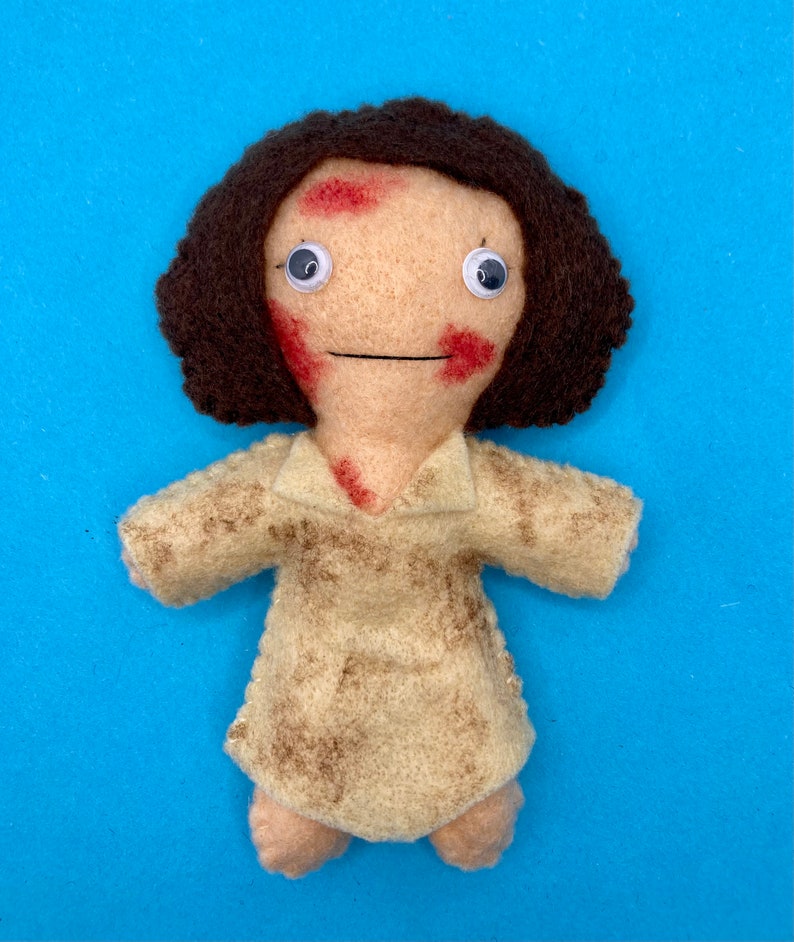 May include: A handmade felt doll with a light tan face, brown hair, and a light tan dress. The doll has red markings on its face and chest, and googly eyes. The doll is set against a blue background.